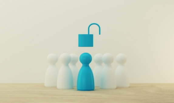 A group of five white game pieces surrounds a single blue game piece on a wooden surface. An open padlock icon hovers above the blue piece, symbolizing unlocked potential or access. The background is plain and neutral.